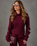 Woman in burgundy cutout-sleeve turtleneck sweater, satin wide-leg pants, aviator sunglasses and woven maroon crossbody bag