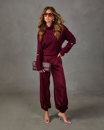 Woman in burgundy cutout-sleeve turtleneck sweater, satin jogger pants, woven clutch, aviator sunglasses and clear heeled sandals