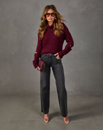 Woman in burgundy cutout-sleeve turtleneck sweater, gray wide-leg jeans, clear heels, and a woven maroon clutch