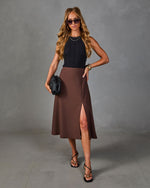Woman wearing black ribbed sleeveless top, brown midi skirt with front slit, black strappy heels, and woven clutch