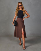 Woman wearing black ribbed sleeveless top, brown midi skirt with front slit, black strappy heels, and woven clutch