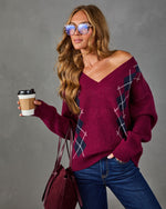 A woman wearing a burgundy off-the-shoulder sweater with a navy and white argyle pattern, paired with blue jeans and holding a coffee cup against a gray studio background.