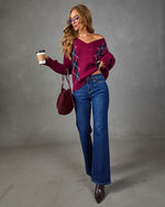 A woman models an off-the-shoulder burgundy sweater with a blue and white argyle pattern on the front, paired with blue jeans, brown boots, and a matching burgundy handbag, holding a coffee cup against a gray studio background.