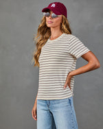 Woman wearing cream striped t-shirt, burgundy LA baseball cap, round sunglasses, and light wash jeans, hand on hip