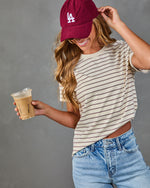 Woman in cream striped crop tee, LA corduroy cap, and light wash jeans holding iced coffee, smiling casually