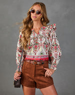 A woman modeling a bohemian-style long-sleeve blouse with a red, green, and blue paisley pattern on an off-white background, paired with brown shorts and sunglasses, against a gray studio backdrop.