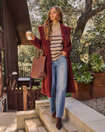 A woman stands on outdoor stone steps in front of a wooden house, wearing a long burgundy fuzzy coat over a beige and navy striped sweater, blue jeans, and brown ankle boots. She holds a coffee cup and a brown woven tote bag.