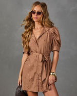 A woman models a brown short puff-sleeve mini dress with a tie waist and subtle tonal pattern, styled with sunglasses, gold jewelry, and a clutch against a gray studio background.