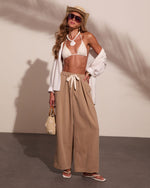 Woman in white bikini top and beige wide-leg pants with straw hat, white cardigan, and woven bag in summer beach style outfit