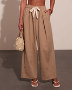 Woman wearing beige linen wide-leg pants with tie waist and pockets, holding woven straw handbag, white sandals