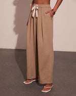Woman wearing beige wide-leg palazzo pants with tie waist and side pockets, paired with white sandals and gold bracelets