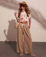 Woman in summer beach outfit wearing white seashell print crop top, tan wide-leg pants, straw hat and sunglasses with woven bag
