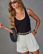 Woman wearing black knit tank top with cream trim, white tailored shorts, black belt, woven leather bag, and oval sunglasses