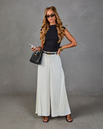 Woman in black sleeveless top, white pleated wide-leg pants, black belt, mini bag, and sandals, posing in studio
