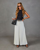 Woman in black sleeveless top, white pleated wide-leg pants, black belt, mini bag, and sandals, posing in studio
