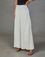White pleated wide-leg trousers with belt loops, worn with black strappy sandals and gold bangles, styled against gray background
