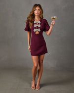 Burgundy puff-sleeve mini dress with rhinestone bow cutouts, styled with nude heels and a champagne coupe
