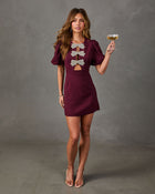 Burgundy puff-sleeve mini dress with rhinestone bow cutouts, styled with nude heels and a champagne coupe