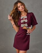 Woman in a burgundy mini dress with puff sleeves and rhinestone bow cutouts, holding a champagne coupe against a gray backdrop
