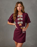 Burgundy puff-sleeve mini dress with rhinestone bow cutout details, styled with a coupe glass against a gray backdrop