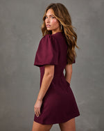 Woman in a burgundy puff sleeve mini dress with a V-neckline, shown from behind against a gray backdrop, glancing over her shoulder