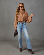 A woman modeling a brown long-sleeve blouse with dotted embroidery, paired with light-wash blue jeans, brown ankle boots, and holding a dark clutch against a gray studio background.
