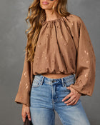 A woman wearing a brown long-sleeve blouse with a subtle tonal pattern and blue jeans, standing against a textured gray studio background.