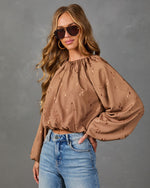 A woman modeling a brown long-sleeve blouse with a subtle tonal pattern, paired with blue jeans, against a textured gray studio background.