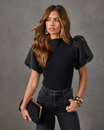 Woman wearing a black knit top with sheer organza puff sleeves, black jeans, gold cuff bracelets and a black clutch