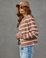 A woman wearing a brown and beige horizontally striped fuzzy cardigan, a matching brown baseball cap, sunglasses, and light blue jeans, posed against a solid gray studio background.