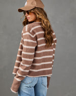 A woman wearing a brown and cream striped fuzzy knit sweater, a tan baseball cap, and light blue jeans, photographed against a textured gray studio background.