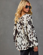 Women's cream and brown cow print long sleeve blouse with balloon sleeves, worn with dark jeans and aviator sunglasses