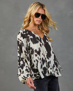 Woman wearing white and black cow print long-sleeve blouse with V-neck, paired with dark jeans and aviator sunglasses