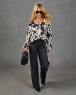 Woman wearing white and black cow print blouse with wide-leg dark wash jeans, black woven bag, sunglasses, and black heeled boots.