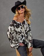 Woman wearing cow print off-shoulder blouse, black cowboy hat, aviator sunglasses, and dark jeans in studio setting