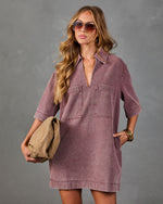 A woman modeling a dusty mauve denim shirt dress with a V-neck, short sleeves, and two chest pockets, holding a tan clutch against a gray studio background.