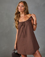 Brown plaid mini dress with ruched neckline and tie detail, modeled with gold jewelry and woven leather bag