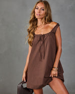 Woman wearing brown gingham mini dress with ruffle neckline, gold bangles, and woven leather bag against gray background
