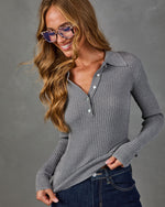 A woman wearing a gray ribbed long-sleeve polo-style knit top with a button placket, paired with dark blue jeans, against a neutral gray studio background.