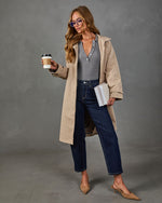 A woman models a beige trench coat over a gray striped top, dark blue jeans, and tan slingback heels, holding a coffee cup and a notebook against a gray studio backdrop.