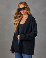 A woman modeling an oversized dark navy open-front cardigan over a black top and blue jeans, photographed against a textured gray studio background.