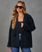 A woman modeling an oversized dark charcoal knit cardigan with front patch pockets and a single visible gold-tone button, worn over a black top and blue jeans against a textured gray studio background.