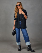 A woman models a black double-breasted blazer over a black top, paired with blue cuffed jeans, black ankle boots, and sunglasses, holding a black quilted clutch against a gray studio background.