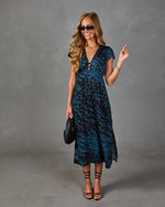 Woman wearing blue leopard print midi dress with V-neckline, cap sleeves, black strappy heels, sunglasses, and woven clutch