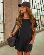 Woman wearing black short-sleeve romper with patch pockets, dark quilted tote bag, and charcoal baseball cap, auburn wavy hair