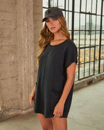 Woman wearing black mini t-shirt dress with front pockets and LA baseball cap, standing in urban loft setting