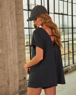 Woman modeling black open-back romper with cross-strap detail, wearing charcoal baseball cap and gold hoop earrings, industrial loft setting
