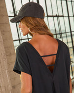 Woman wearing black open-back V-neck top with strappy detail, dark baseball cap, gold hoop earrings, auburn hair