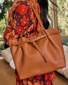 A person wearing a floral dress carries a large tan pebbled leather tote bag with a strap and front buckle detail in a bright indoor setting.