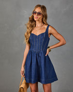 Dark wash denim mini dress with corset bodice and flared skirt, styled with aviator sunglasses and tan handbag